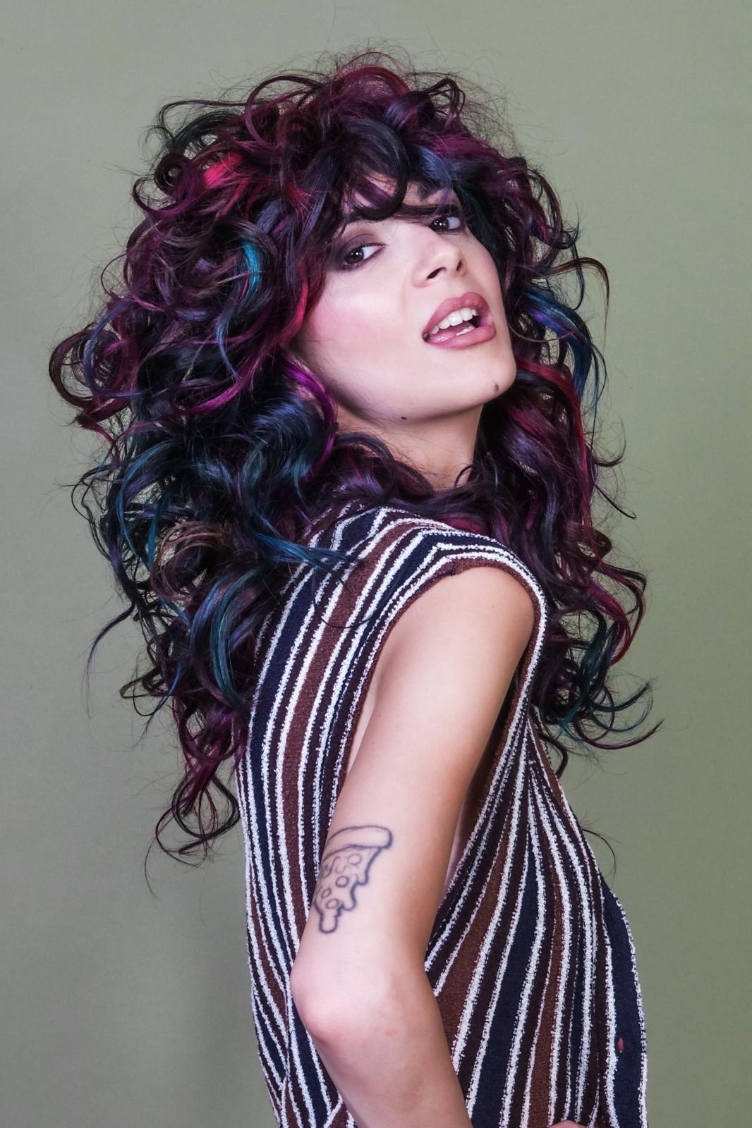 Una chioma lunga e voluminosa, costruita su ricci morbidi e dinamici che creano movimento e carattere. La base scura è attraversata da accenti viola, fucsia e blu, per un risultato vibrante e creativo. Long, voluminous hair shaped by soft, dynamic curls that create movement and character. The dark base is illuminated by violet, fuchsia and blue accents for a vibrant, creative finish.